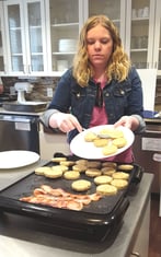 Kristin pushes frozen sausage patties off a plate and onto an electric griddle to sizzle alongside the bacon.