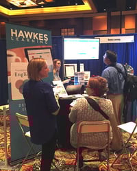 Two Hawkes representatives chat with two instructors in front of the Hawkes Learning booth as the TV monitor displaying the courseware bathes the room in an ethereal fluorescent glow.