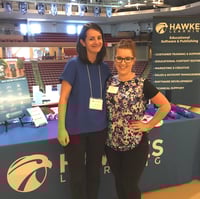 Florie and Megan stand on stage in front of a table with the Hawkes Learning logo.