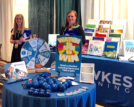 Ashlyn and Liz stand behind a table full of giveaways like yo-yos, buttons, and stress balls, as well as a spinning wheel for prizes.