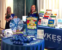 Ashlyn and Liz stand behind a table full of giveaways like yo-yos, buttons, and stress balls, as well as a spinning wheel for prizes.