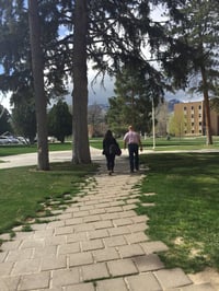 Ashley and Professor Potter walk away from the camera along a wooded path on campus.