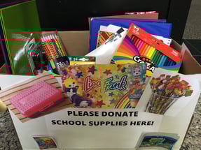 A box full of folders, binders, pencils, and colored pencils. The box has a sign that says 'Please donate school supplies here.'