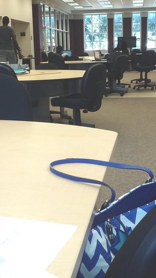 Several desks with office chairs and computers are in a well-lit room with large windows on the back wall.
