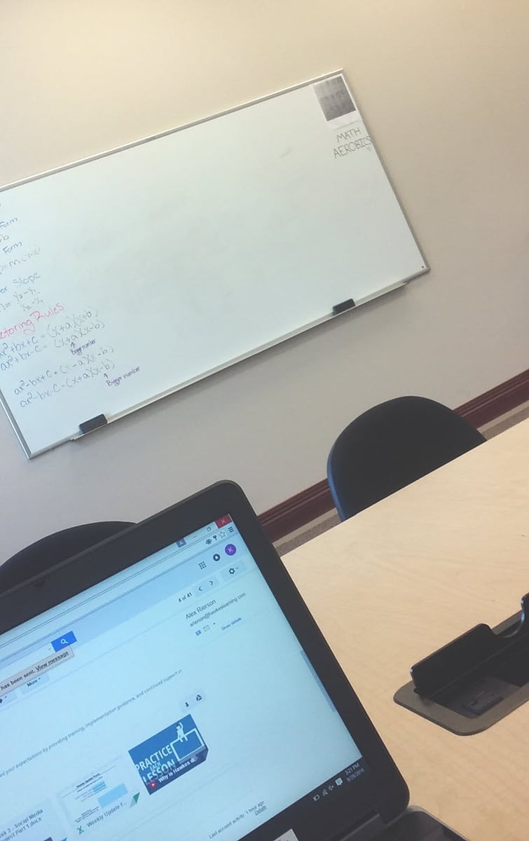 A laptop screen is in the foreground; beyond is a whiteboard with equations written on it.
