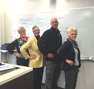 Instructors standing in front of a classroom white board strike the Hawkes pose by putting their hands on hips.